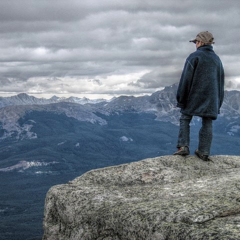 A boy stands at the edge of a cliff and looks out at a valley below and snow covered peaks in the distance.