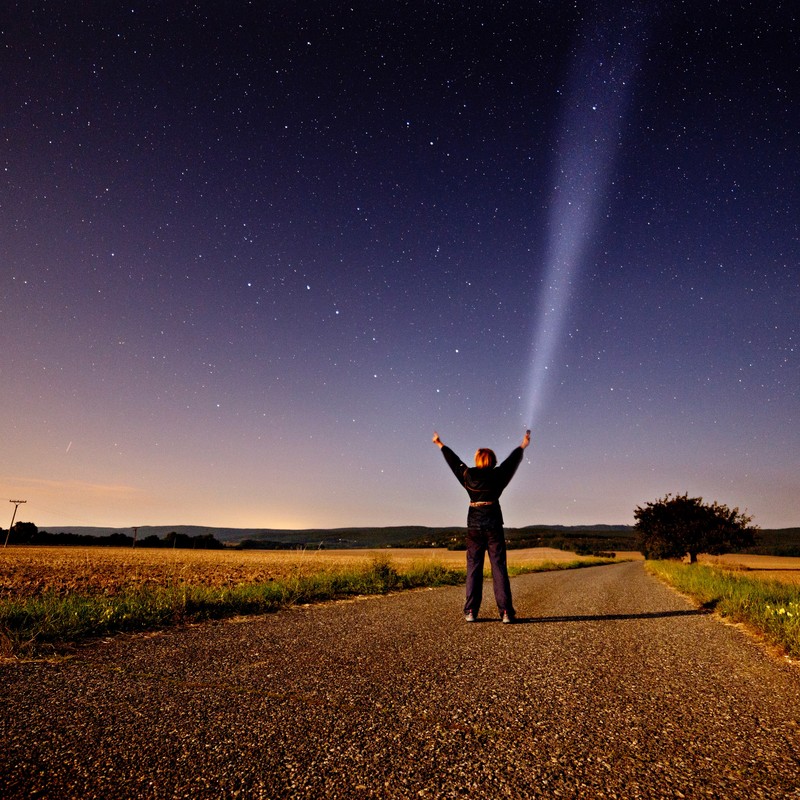 Una mujer se para en medio de un camino rural por la noche y llega hacia el cielo lleno de estrellas arriba.