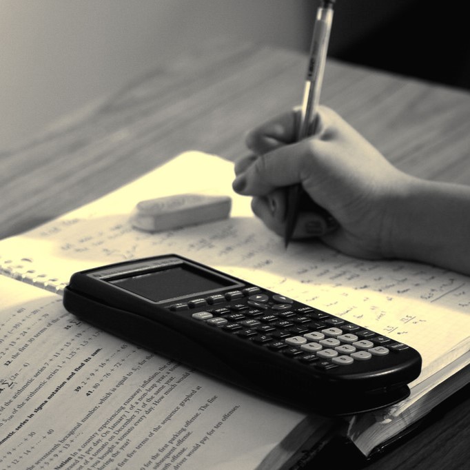 A student works on math homework with an open book, spiral notebook, and calculator. A student works on math homework with an open book, spiral notebook, and calculator.