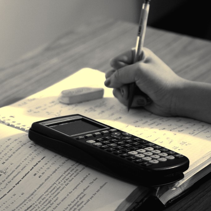 A student works on math homework with an open book, spiral notebook, and calculator.