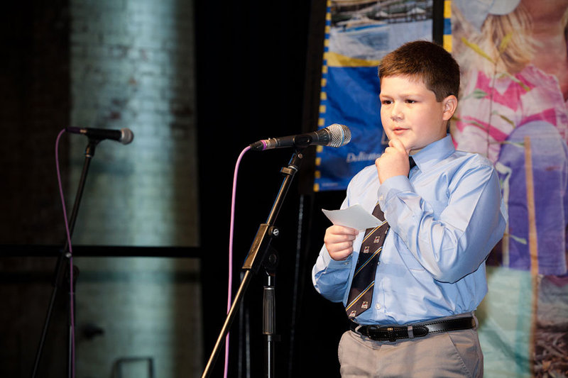 A boy holding a note card steps up to a microphone to give a public presentation.