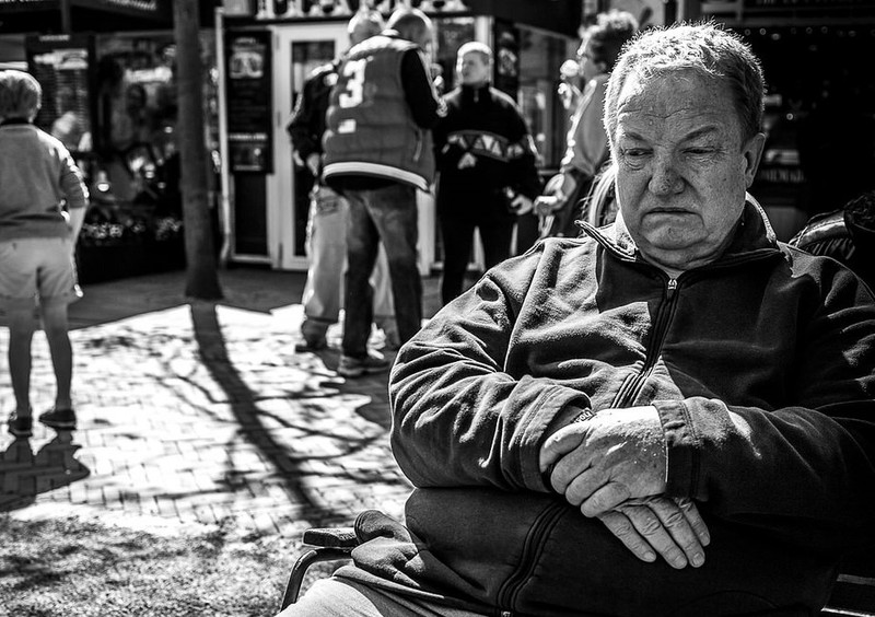 A man sits alone on public bench with his arms crossed and downcast eyes. 