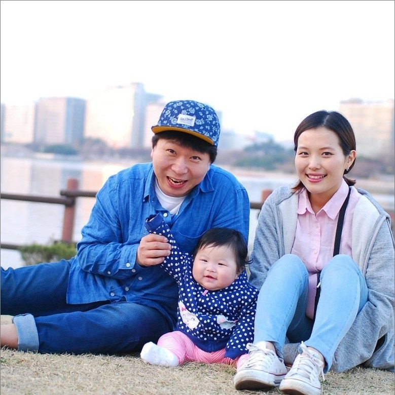 A young mother and father sit with their infant in a park.