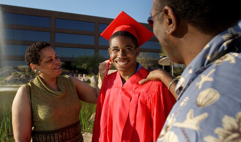 Padres sonrientes se paran con su hijo adulto joven que está vestido con una gorra y bata de graduación.