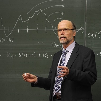 A stereotypical image of a professor - a white, middle-aged man with glasses and a beard, dressed in a coat and tie stands with chalk in hand in front of a blackboard which displays a mathematical formula.
