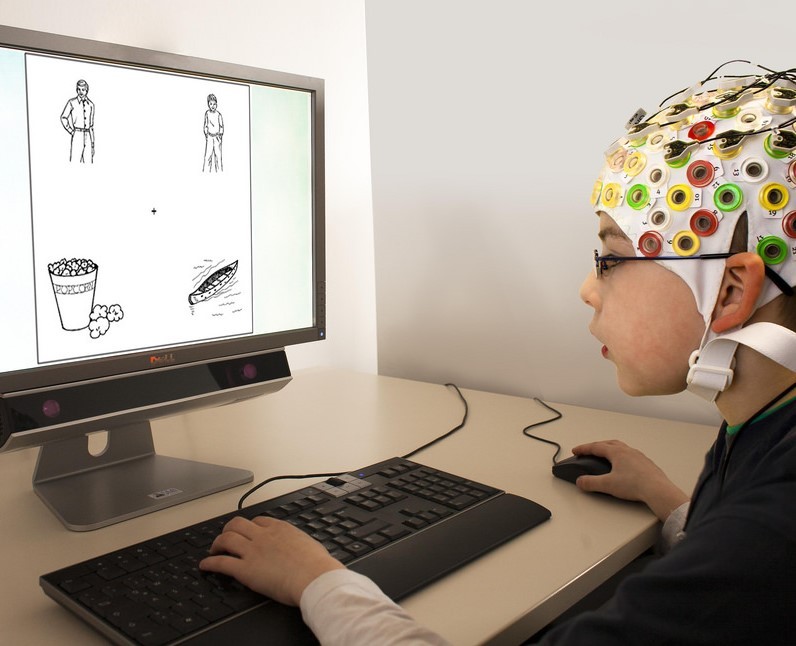 Un niño con gorra EEG usa un mouse y un teclado mientras mira imágenes en un monitor de computadora durante un experimento.