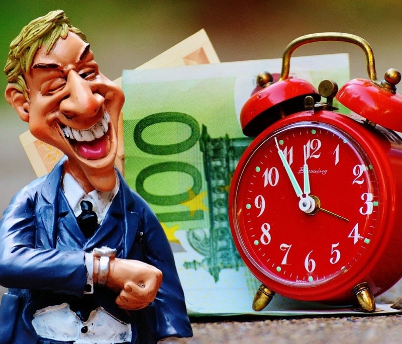 A business man stands grinning and looking at his watch with a clock and some cash visible in the background. 