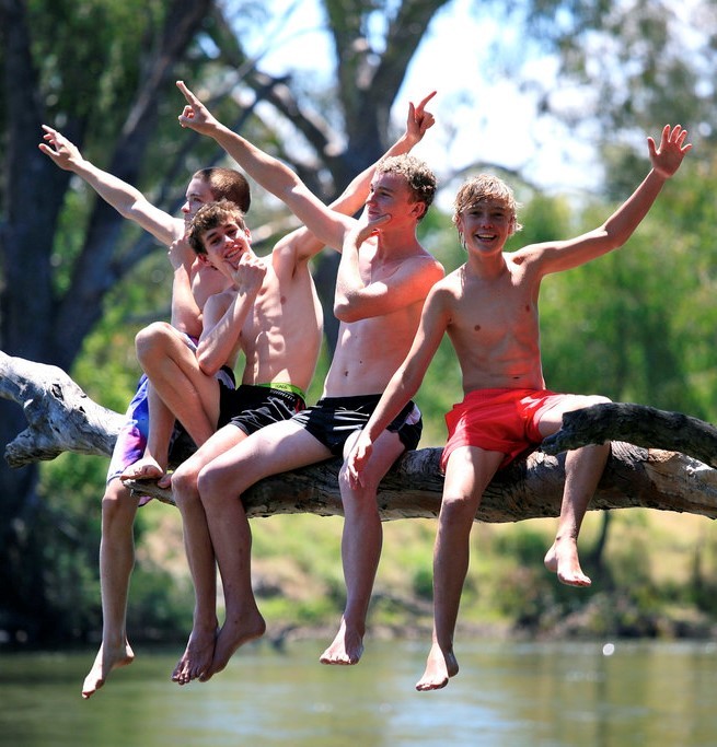 A group of teen aged boys in swimming trunks sit together on a tree branch above a pond as they smile and show off for the camera.