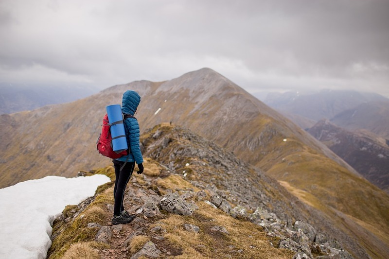 A hiker stands high on a ridge and looks at cloud covered mountains in the distance.