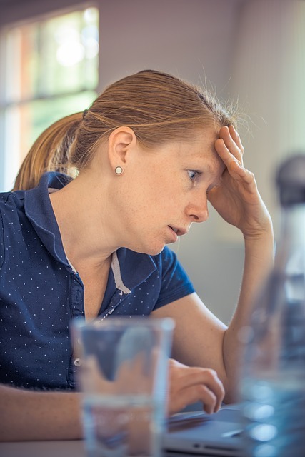 A woman with an overwhelmed expression puts her hand to her forehead as she looks at a computer screen.