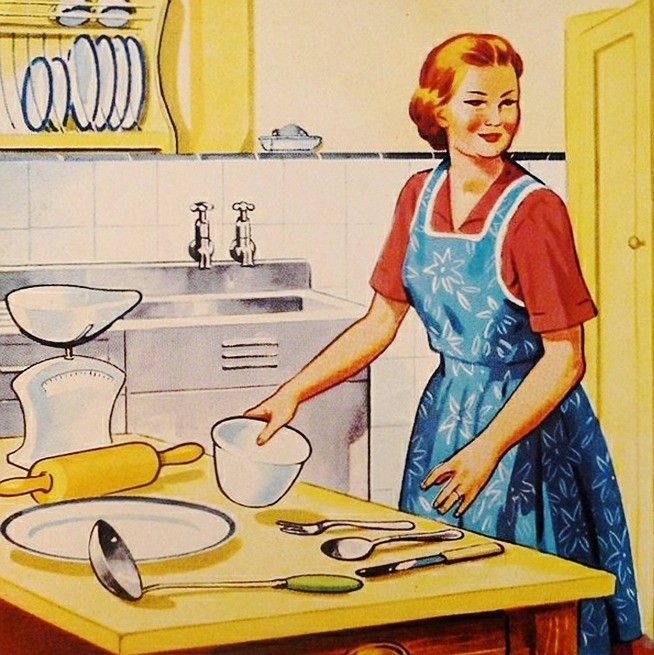 A stereotypical housewife of the 1950s stands in her kitchen wearing an apron with a table full of cooking utensils in front of her.