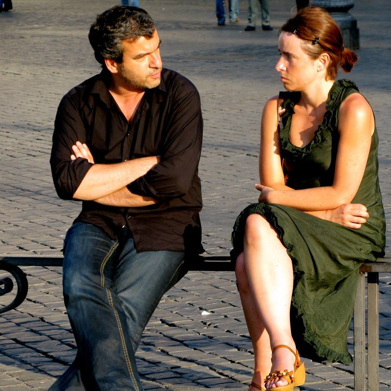 A couple sit next to each other on a public bench with arms and legs crossed in a defensive posture. They stare at each other with frowning faces. 