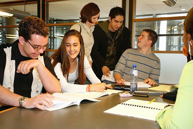 A group of college students sit around a table talking and looking through textbooks and notebooks.