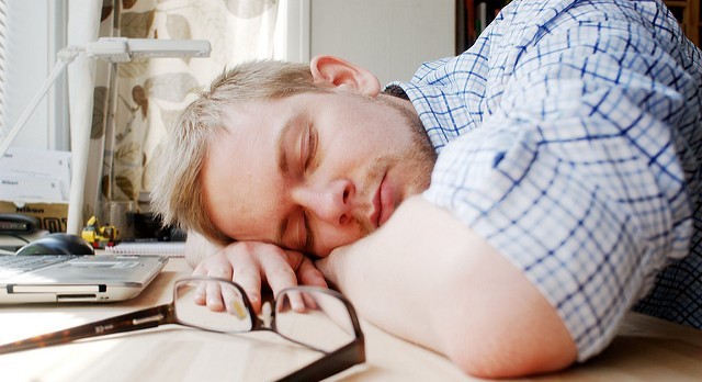 A tired college student sleeps with his head on his desk with his laptop nearby.