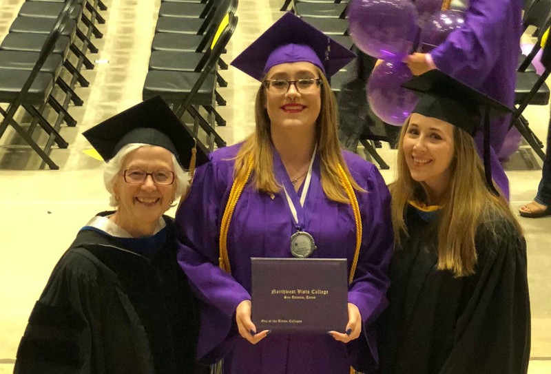 An image of the May 2018 Commencement Ceremony at NW Vista College. Three women in caps and gowns. Two women on each side wearing black cap and gowns and the woman in the middle wearing a purple cap and gown holding a diploma.