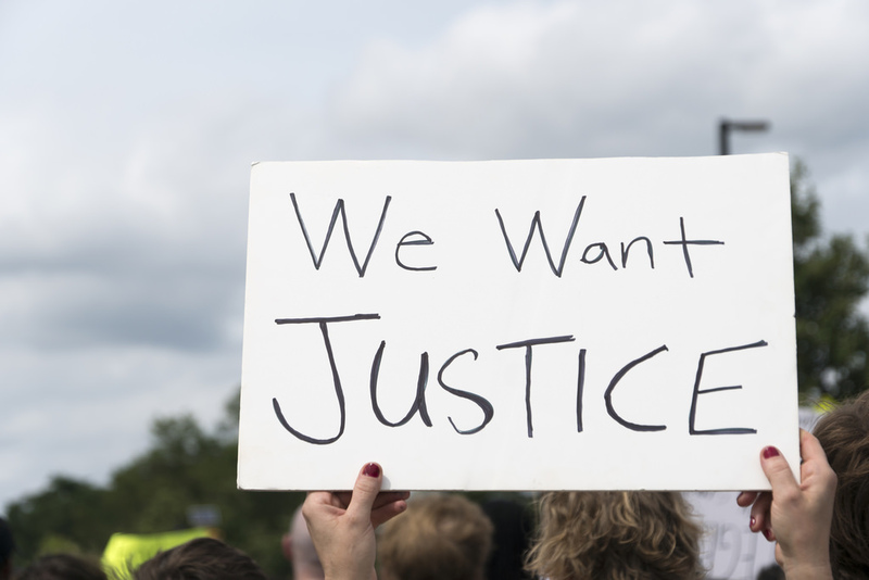 This image depicts two hands holding a sign high. The sign is white and has a hand-written message that reads "We want justice" in black letters. In the distance, one can see the fuzzy images of many heads, suggesting that this sign is being held up at a gathering such as a protest. There are trees in the distance. 