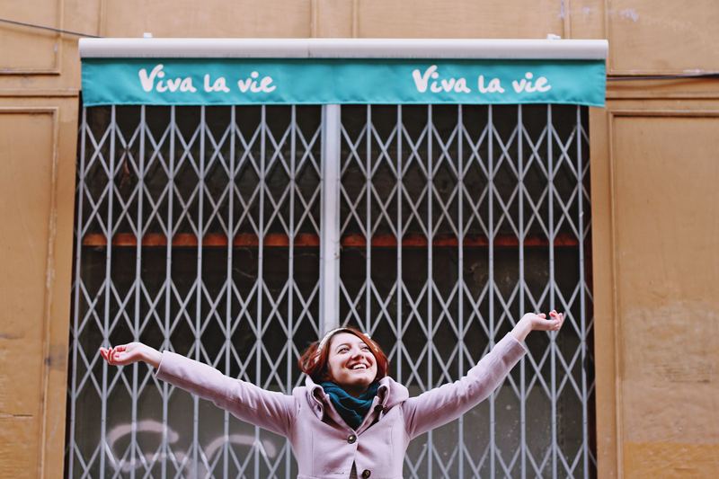 A picture of a woman smiling with her arms raised, standing in front of a storefront with a banner that reads "Viva la vie"