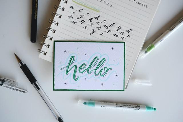 A notepad with a word "Hello" on it on top of another notepad that has the alphabet on it, various pens laying throughout 